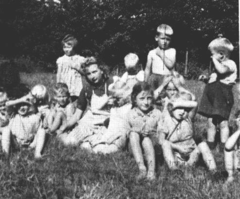 Die Spielwiese fr den Kindergarten befand sich auf der anderen Straenseite auf einer Weide am Gaster Weg. Die Kindertante Emma Bruns war nur kurze Zeit dabei. Links neben ihr steht Thea Remmers, davor sitzt Berthold Siebrands, und das Mdchen mit dem gesenkten Kopf knnte Henni Lhring sein. Wer sich da mit dem Unterarm vor der Sonne schtzt, wissen wir nicht, aber ganz links sitzt noch Berta Penning. Der dunkle Fleck ber ihr verdeckt die Kindertante Wilhelmine Hahn, die sich "weggezaubert" hat. Rechts neben Tante Emma das Mdchen mit der Hand vor den Augen knnte Beate Bruns sein, der Junge dahinter mit dem gesenkten Kopf heit Gnter Meyerhoff. Neben ihm steht sein groer Bruder Gerd, und vor dem sitzt mit den dunklen Haaren Karla Poelmann. Das Mdchen mit den beiden Hnden am Kopf wollte ihren Namen nicht nennen, aber vor ihr sitzt mit dem Arm vor den Augen Helga Poelmann. Der stehende Junge rechts ist Heinz Schmidt.