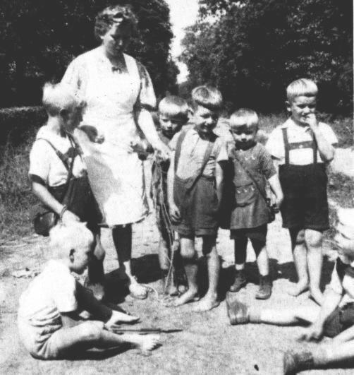 Die Kindertante nne bespricht mit den Jungen ein neues Spiel im Sandkasten. Links sitzt Oswald Steenblock und neben ihm steht Reinhold Brinkmann mit seiner umgehngten Tasche, in der das Frhstcksbrot war. Rechts von Tante nne stehen die Zwillinge Johann und Volkmar Boekhoff und vor ihnen Fritz Kuhlemann. Auch Johann Duis ist auf "leloot Foeten". Neben ihm sitzt am Rande Helmut Schmidt. Die Fe wurden spter im Saal auf der Bhne geschrubbt, bis sie wieder sauber waren. Morgens, mittags und abends wurden die Kinder von den Tanten geholt und wieder weggebracht - zu Fu oder mit dem Rad, nicht mit dem Bus! -