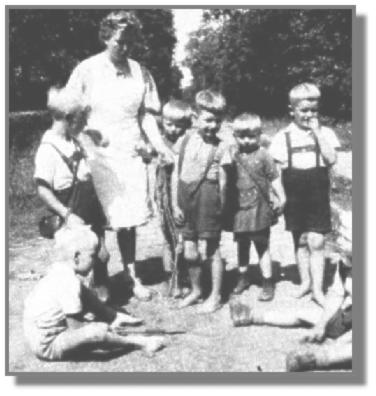 Die Kindertante nne bespricht mit den Jungen ein neues Spiel im Sandkasten. Links sitzt Oswald Steenblock und neben ihm steht Reinhold Brinkmann mit seiner umgehngten Tasche, in der das Frhstcksbrot war. Rechts von Tante nne stehen die Zwillinge Johann und Volkmar Boekhoff und vor ihnen Fritz Kuhlemann. Auch Johann Duis ist auf "leloot Foeten". Neben ihm sitzt am Rande Helmut Schmidt. Die Fe wurden spter im Saal auf der Bhne geschrubbt, bis sie wieder sauber waren. Morgens, mittags und abends wurden die Kinder von den Tanten geholt und wieder weggebracht - zu Fu oder mit dem Rad, nicht mit dem Bus! -